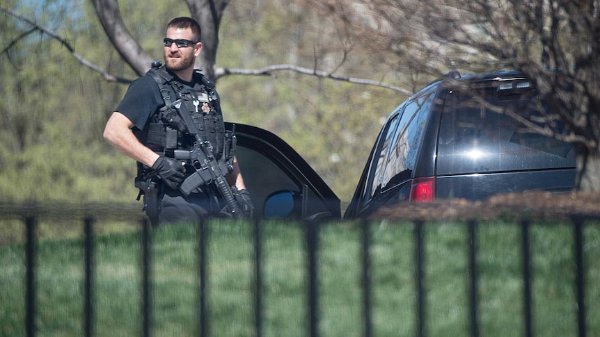 Photo across a fence and lawn to a Secret Service agent, dressed in all black, with a gun standing next to two cars.