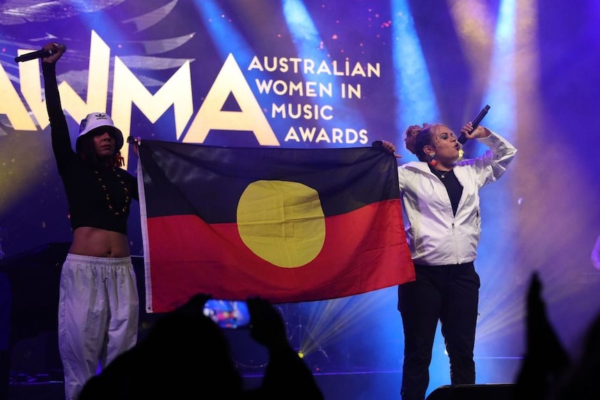 Изображение на Dizzy Doolan и Barkaa, развяващи флаг на аборигените на сцената на Австралийските женски музикални награди