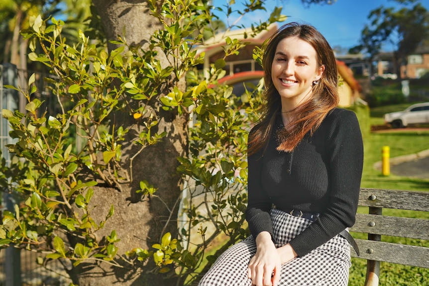 A smiling woman sits outside on a bench.