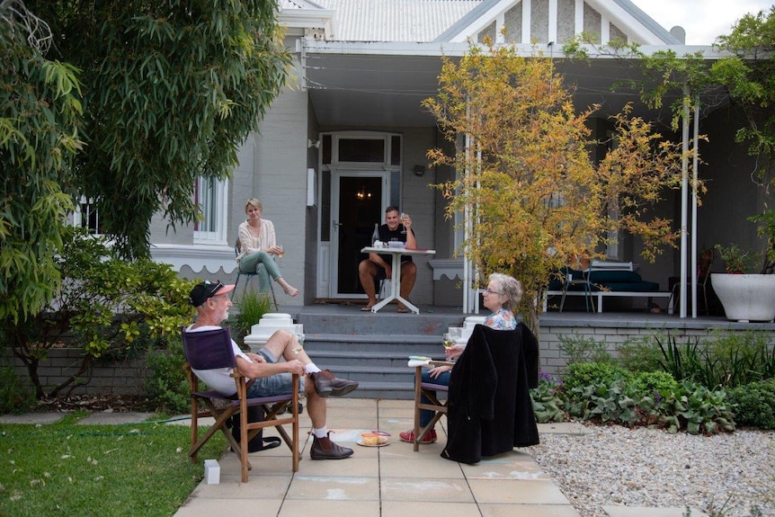 Four people sit, with drinks, distanced from each other, on veranda and front yard