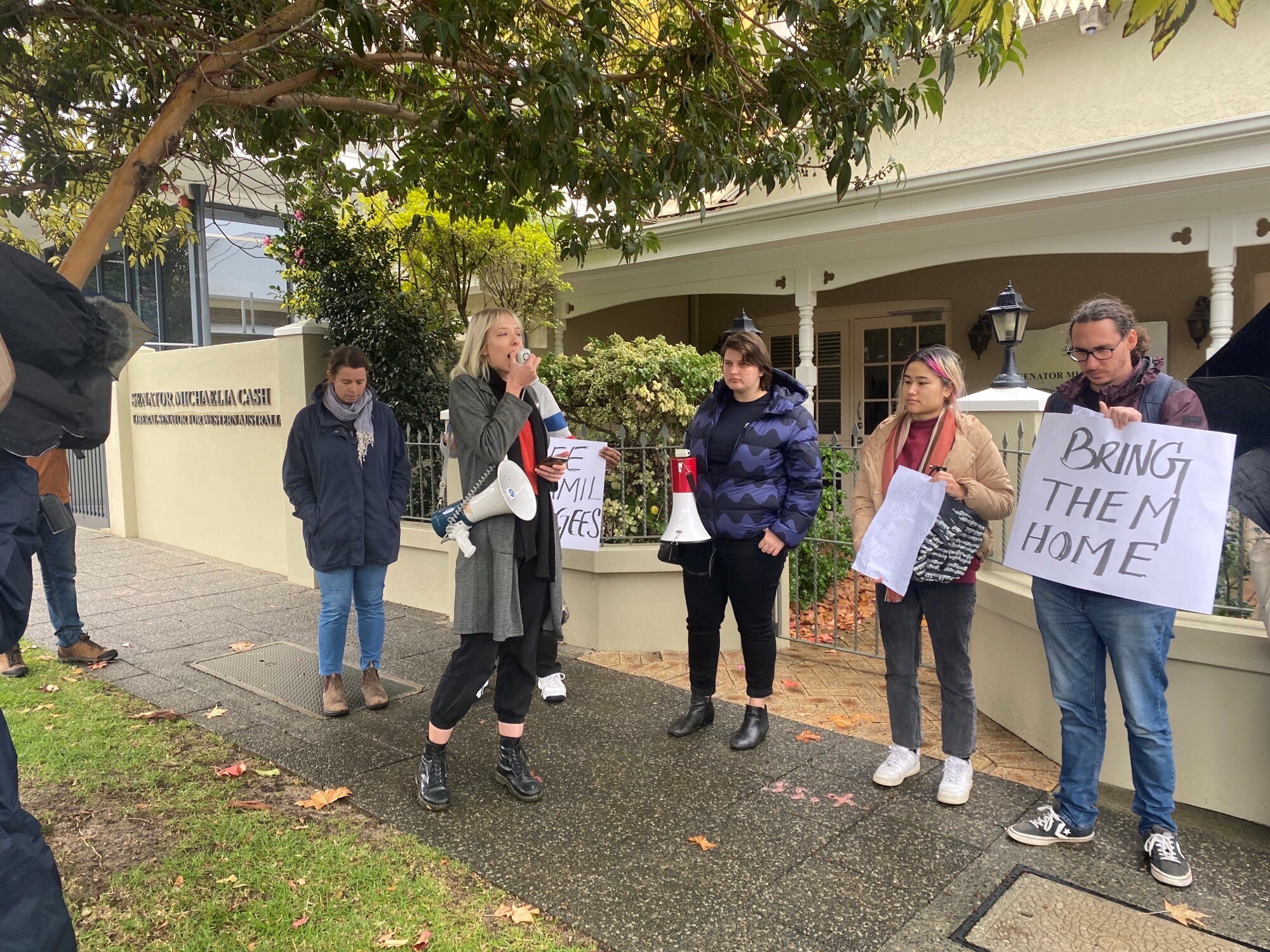 A woman speaks into a loudspeaker, four others hold signs including one saying 'bring them home'.