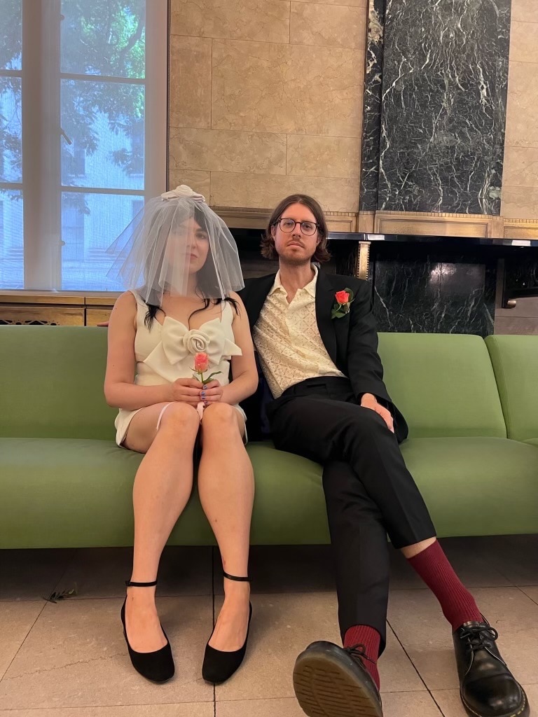Emily Barclay, left, wears a white wedding dress and veil as she sits on a green sofa next to Tom Ward, wearing a suit. 
