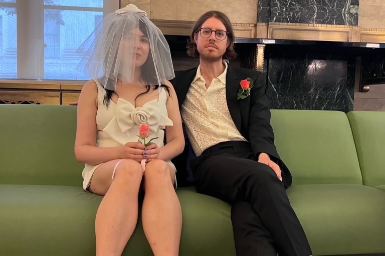 Emily Barclay, left, wears a white wedding dress and veil as she sits on a green sofa next to Tom Ward, wearing a suit. 
