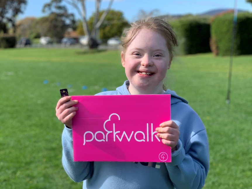 A young girl with blonde hair holds her parkrun certificate and smiles. 