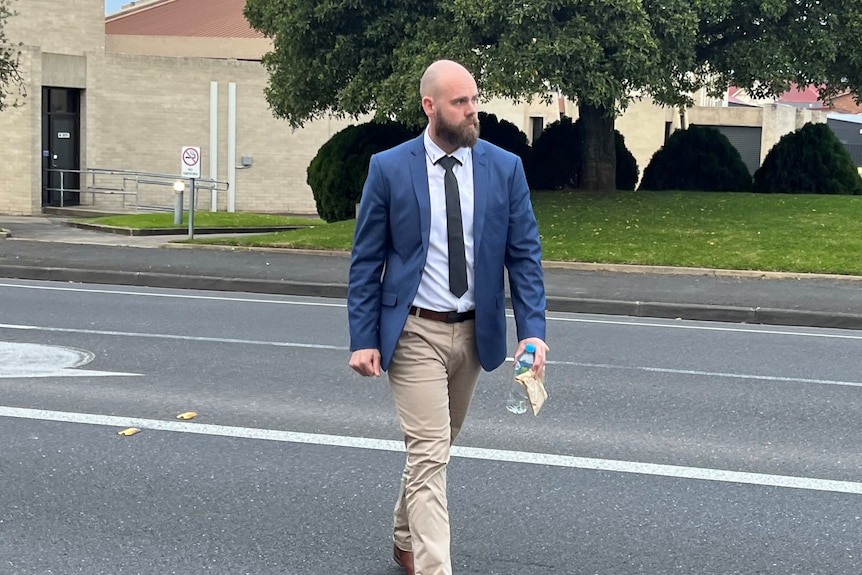 A bald man with a beard wearing a blue blazer, tie and tan pants walks across a street.