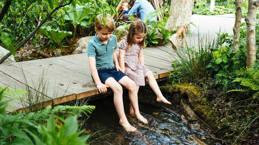 Prince George and Princess Charlotte sit on a wooden bridge and dip their feet in a creek