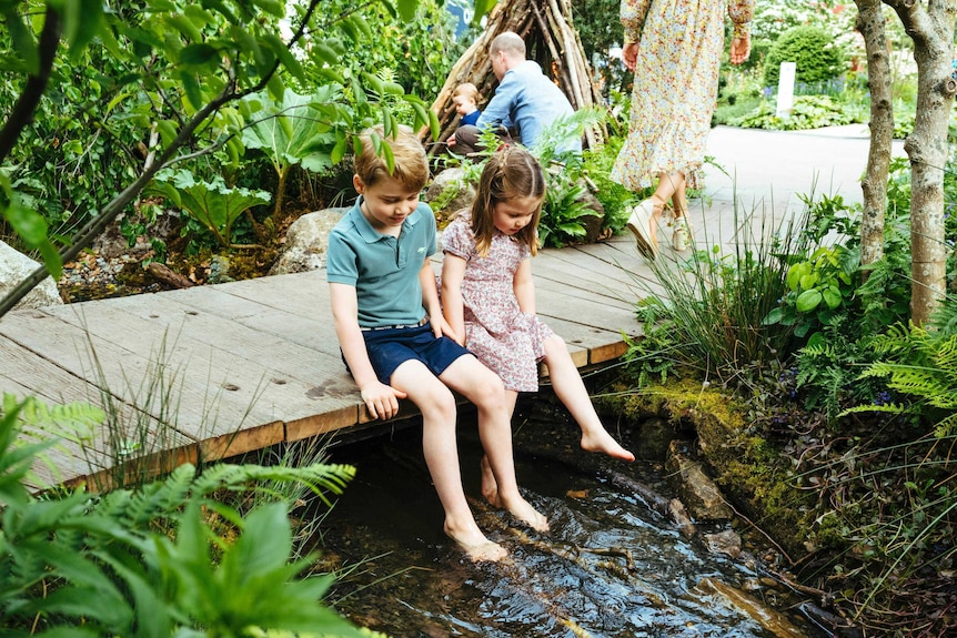 Prince George and Princess Charlotte sit on a wooden bridge and dip their feet in a creek