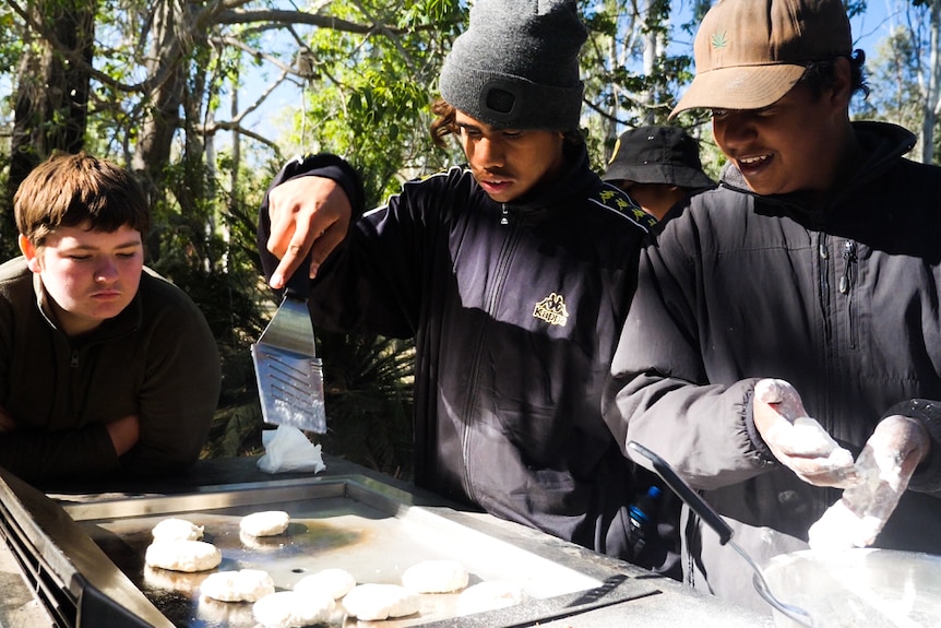 Three boys stand around a barbecue cooking cakes on the grill.