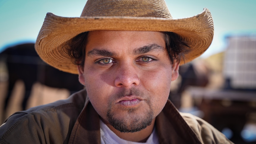 A close-up shot of a young stockman wearing a straw Akubra, staring into the camera lens. 