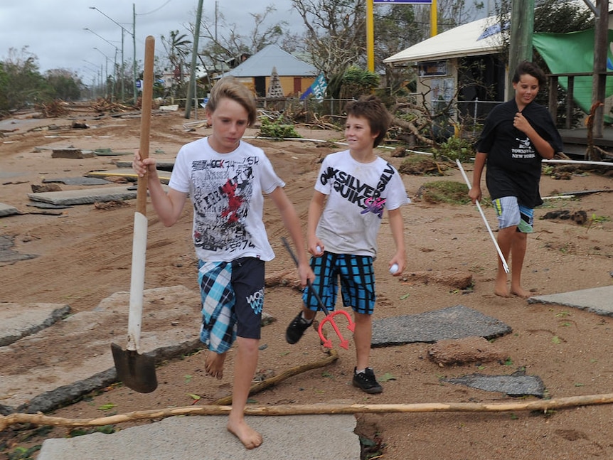 Three boys carrying sticks walk among debris covering a street after a cyclone.