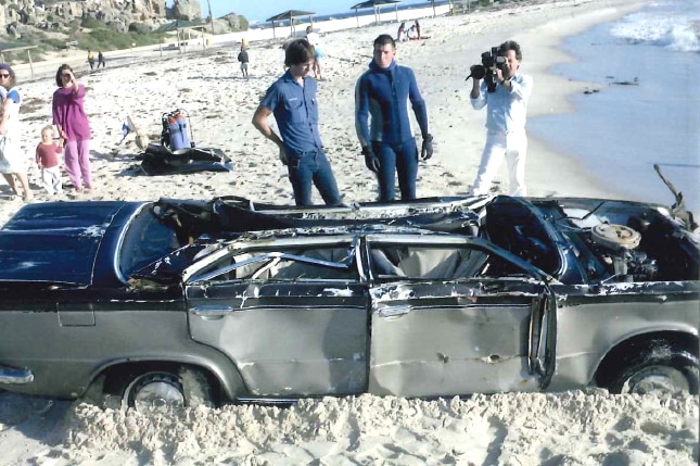 People on a beach stand and look at a badly damaged car, missing a roof and a bonnet.