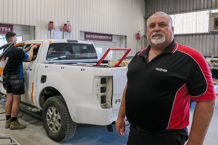 Dave Rogers stands in front of a white ute in the workshop which a man is repairing.