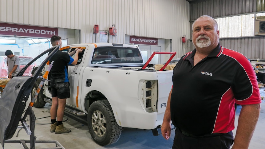 Dave Rogers stands in front of a white ute in the workshop which a man is repairing.