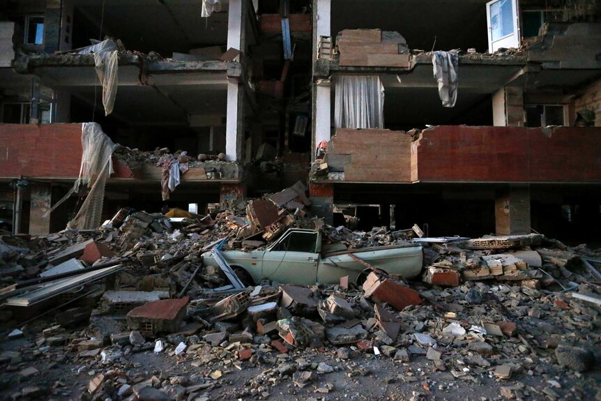 A car sits crushed by debris in front of a damaged building.
