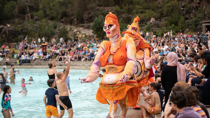 Outdoor shot with 2 large grotesque puppets with orange clothes and hair beside pool with kids swimming and a crowd at the side.