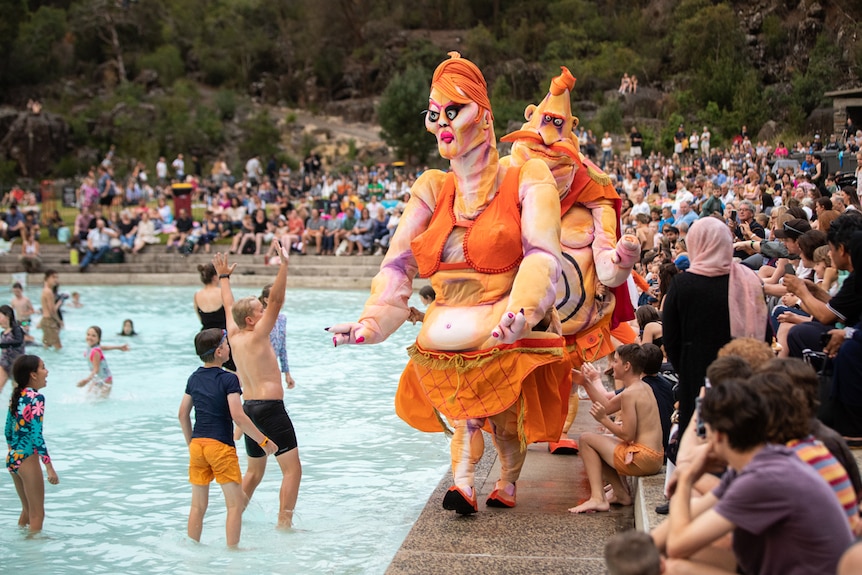 Outdoor shot with 2 large grotesque puppets with orange clothes and hair beside pool with kids swimming and a crowd at the side.