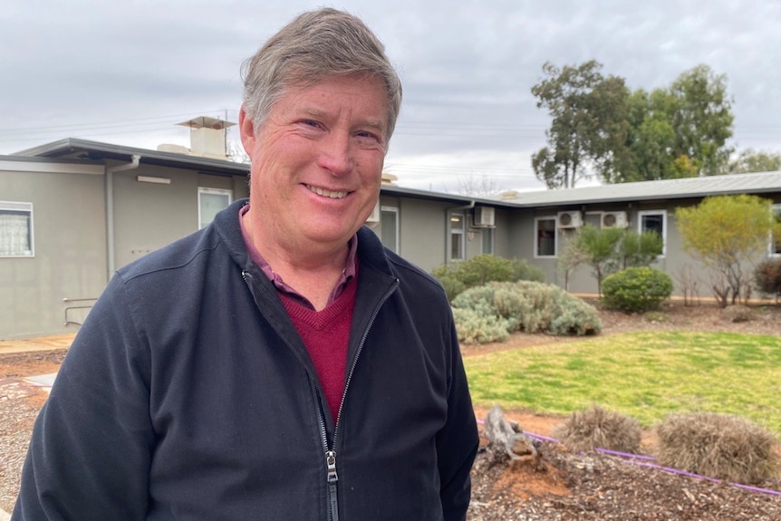 A man smiling at the camera. There is a building and a garden behind him. 