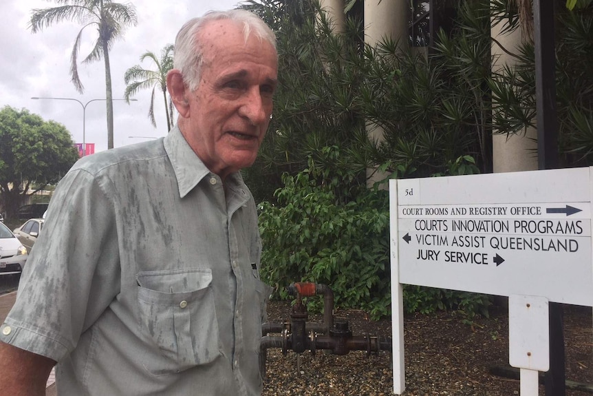 A man with white hair standing to the left of a white sign.