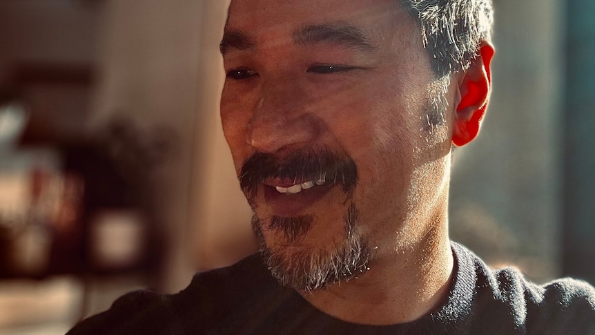 Sun rays fall across the face of a man with short black hair and a goatee who is looking down and gently smiling