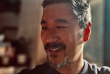 Sun rays fall across the face of a man with short black hair and a goatee who is looking down and gently smiling
