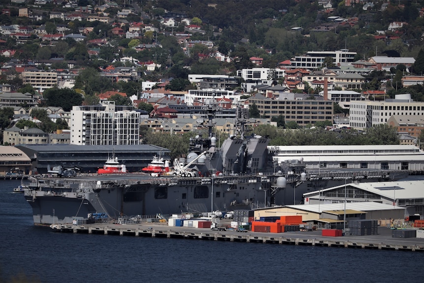 Wide shot of a US warship docked in port with city scape behind.