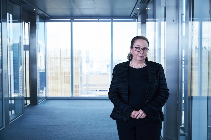 Woman in an office building near the lifts. A glass wall is behind her and she's wearing a formal jacket and skirt. 