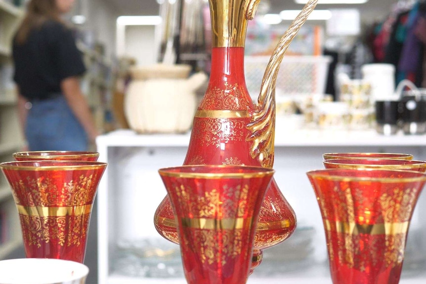 A red and gold jug and matching cups are seen with a person in the background and shelves around them.
