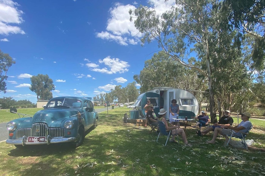 group of people sitting around a table relaxing outside a vintage caravan and car