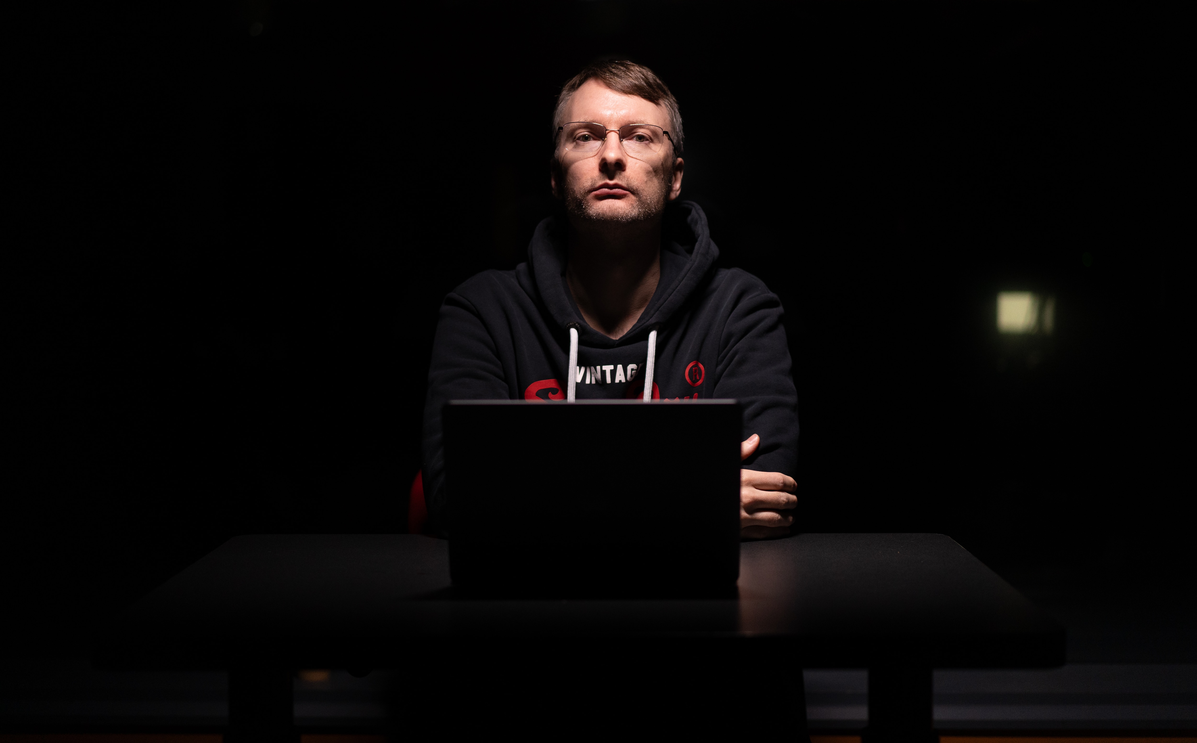 身穿黑色连帽衫的男子坐在黑暗的房间里，面前有一台笔记本电脑。