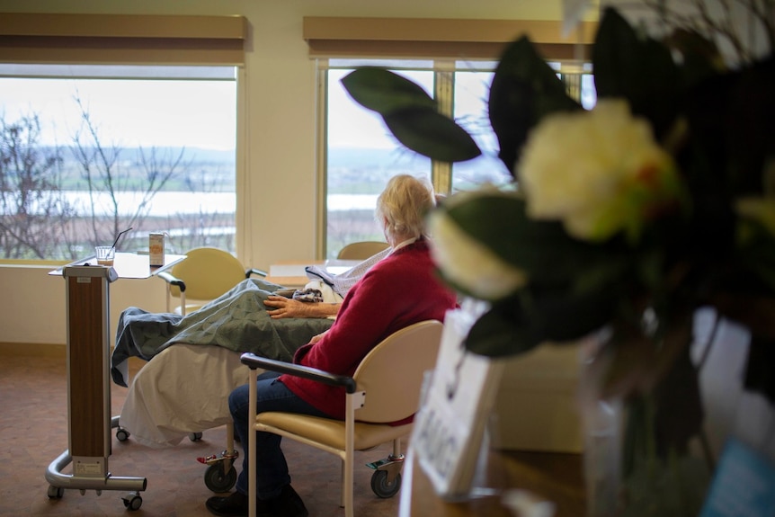 A woman sits next to a resident in an aged care bed