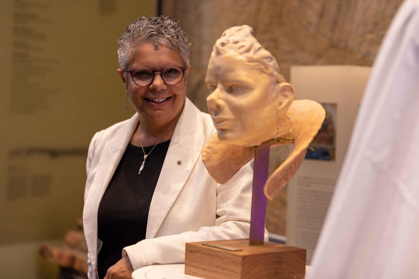 Deborah Cheetham Fraillon stands beside her own bust of her famous face, made by Anna-Wili Highfield. Deborah is smiling widely.