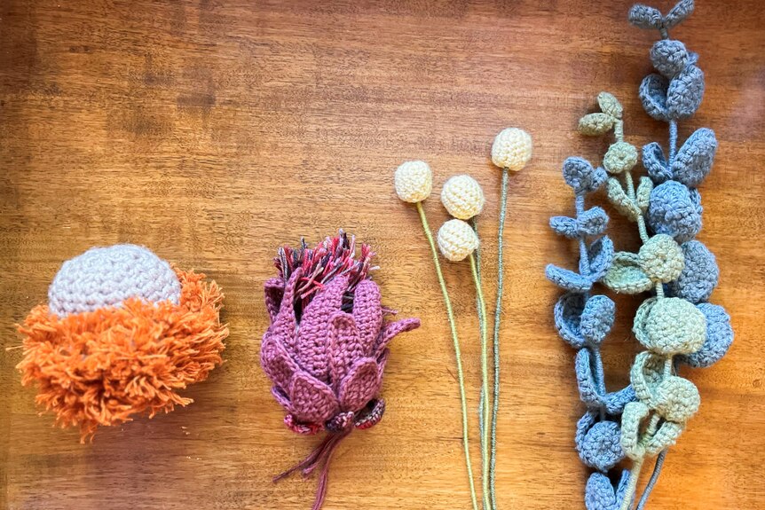 A crochet banksia, princess protea, three billy buttons and three eucalyptus branches on a table.