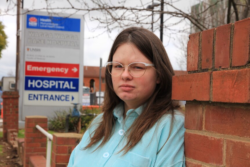 A woman leaning against a brick wall looks sternly at the camera in front of a hospital entrance sign. She has long brown hair. 
