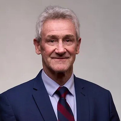 A man with grey hair and a suit looks straight at the camera.