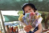 A young girl sits on a boat in a playground, holding pretend money. She is smiling.