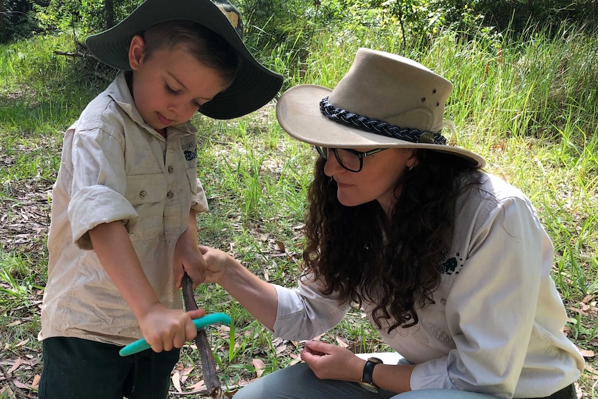 A boy cuts a stick in the bush while a teacher observes.