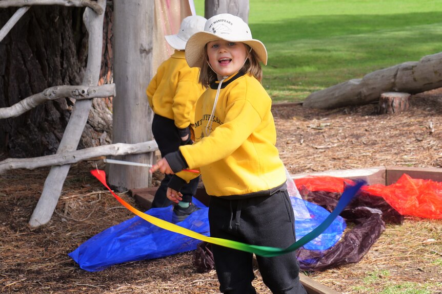 Girl in yellow uniform swings rainbow ribbon around looking at camera.