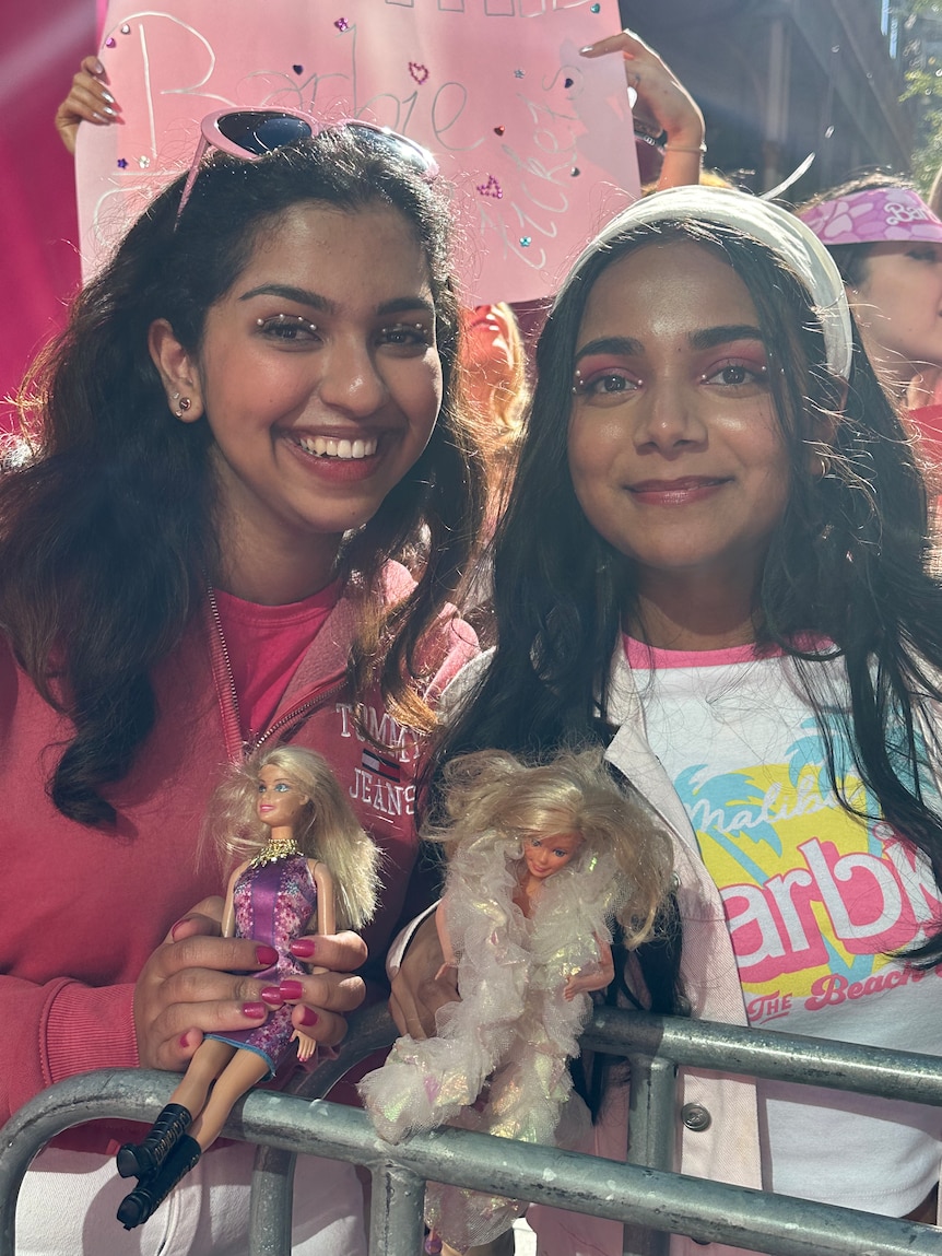 Two young women smiling and holding Barbie dolls wearing Barbie themed clothing