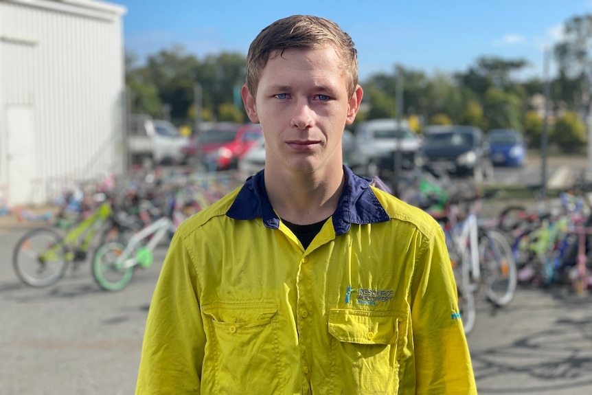 James Holder, brown hair, blue eyes, high vis shirt, looks at the camera with a straight face, bikes behind.