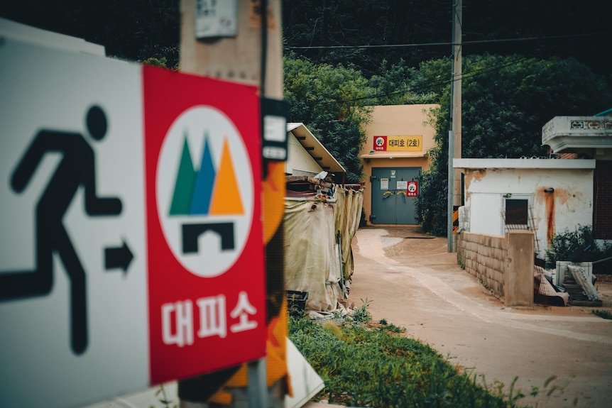 A sign featuring a stick figure running in the foreground with a bomb shelter down the road in the background 