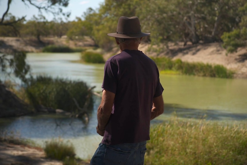 Man in a t-shirt and jeans with a hat looking over a river from behind. 