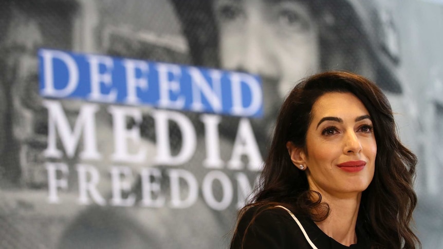 Amal Clooney in front of a sign bearing the words 'defend media freedom' looks to the right of camera