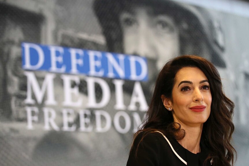 Amal Clooney in front of a sign bearing the words 'defend media freedom' looks to the right of camera