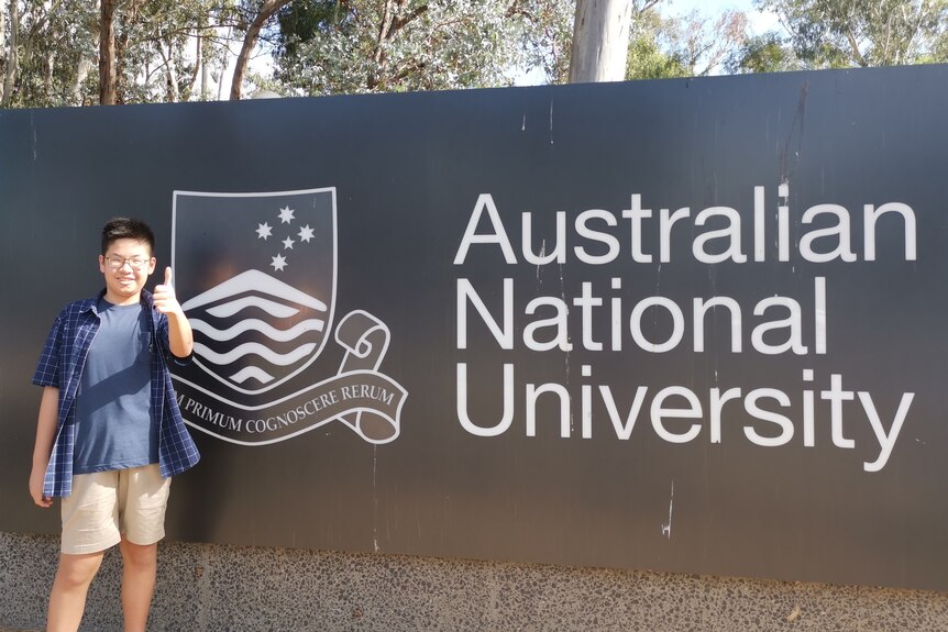 A teenage boy stands in front of an ANU sign.