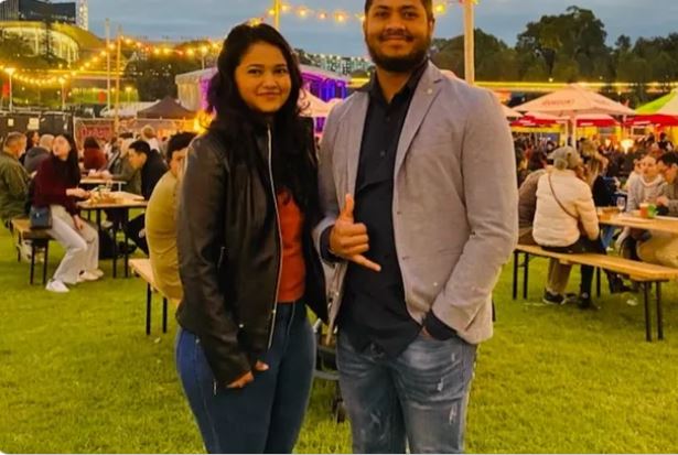 A young women and man stand together smiling at a festival, behind them are people sitting at tables with fairy lights above