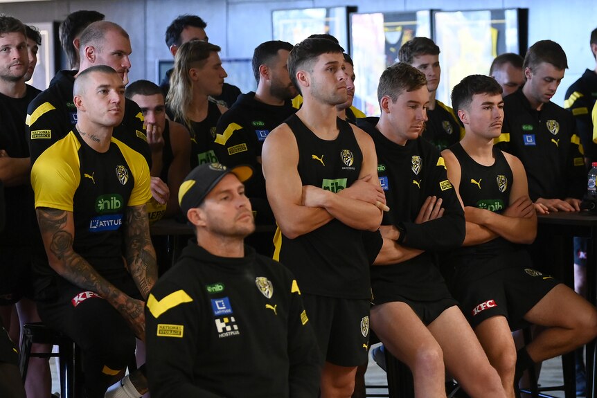 A group of Richmond AFL players sit arms folded, unsmiling, as they look at a press conference taking place. 