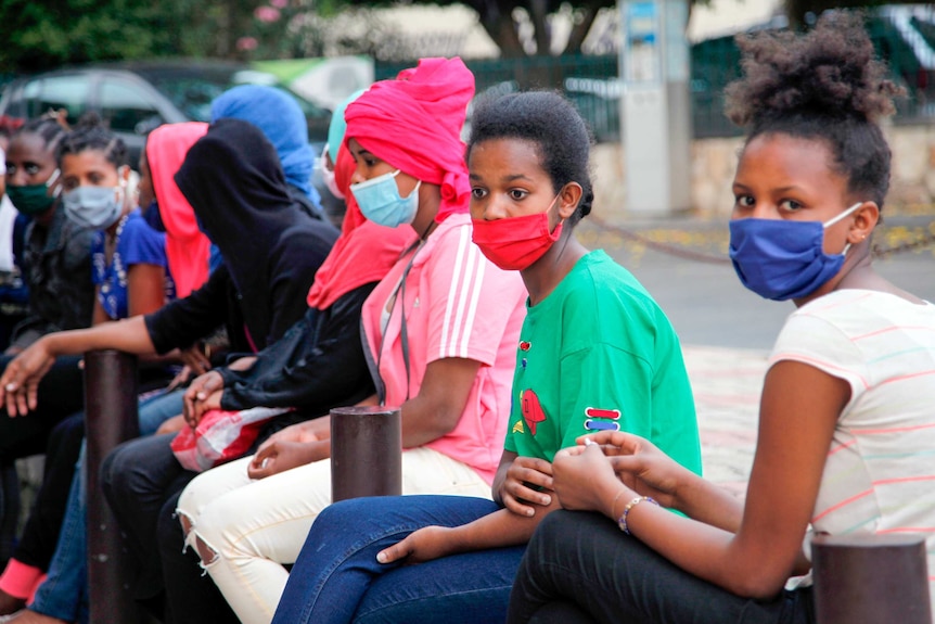 A group of young women in face masks sit on a footpath
