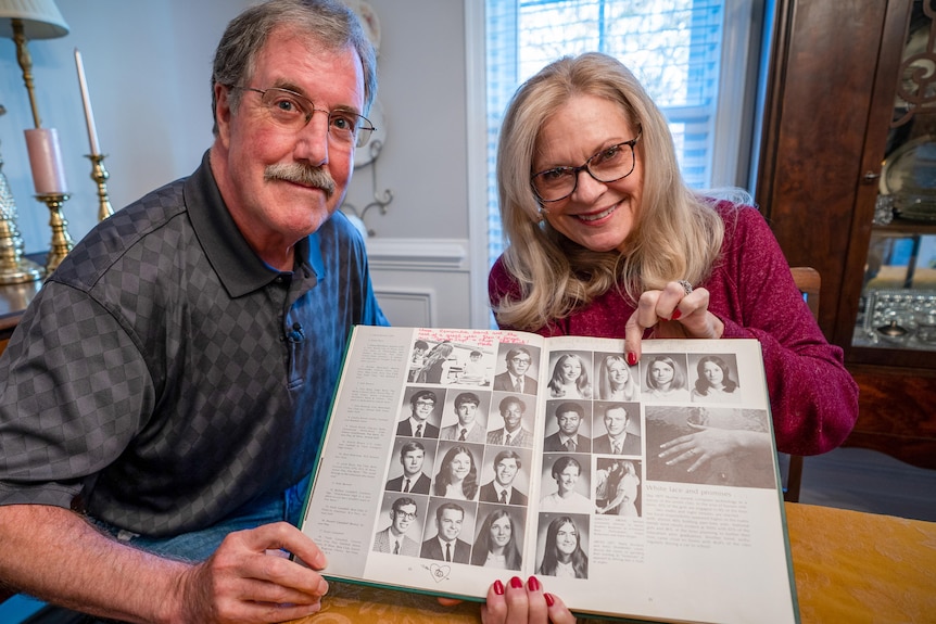 A man and a woman sit at a dining table, holding an old high school year book between them