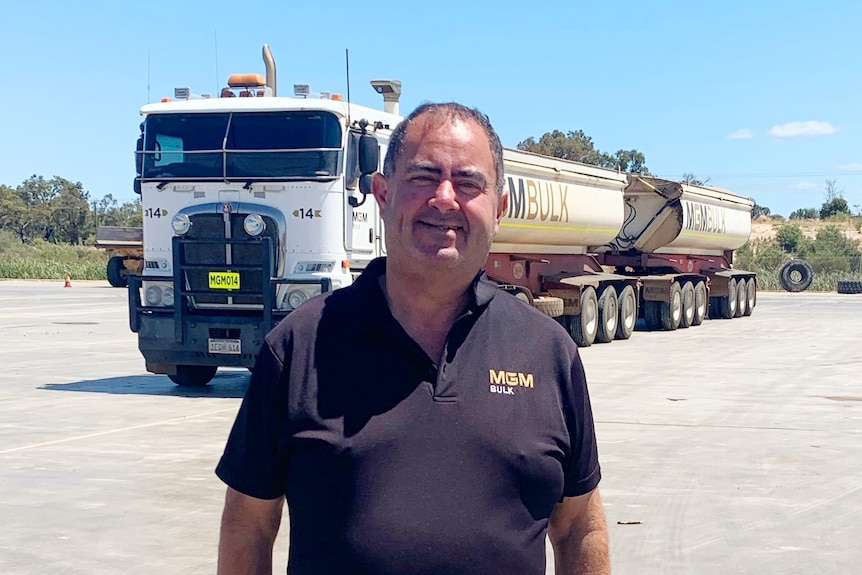 A short-haired man in dark clothes stands in front of a road train on a sunny day.