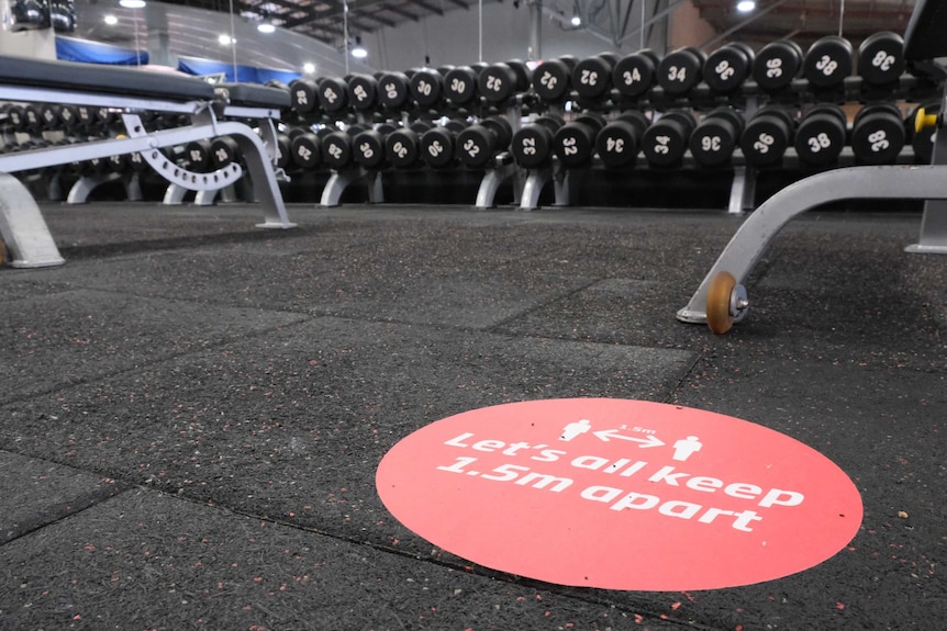 A sticker on the floor reminds patrons to socially distance at a gym, with weights in the background.